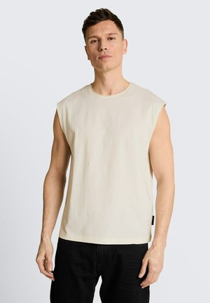 Homme aux cheveux courts et bruns portant une chemise beige sans manches et un pantalon noir, debout devant un fond gris clair.