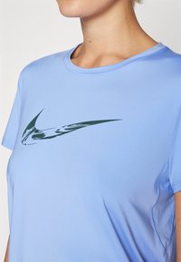Camiseta deportiva azul claro hecha de una tela suave, con un logo de swoosh verde oscuro en el frente, cuello redondo y mangas cortas.