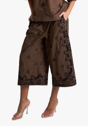 Pantaloni marroni a gamba larga e taglio corto con motivi ricamati neri, indossati con top abbinato e sandali trasparenti con tacco alto.