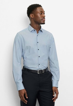 Mann trägt ein hellblau gestreiftes Hemd mit Knöpfen und schwarze Gürtelhose, steht vor einem schlichten weißen Hintergrund und schaut nach rechts.