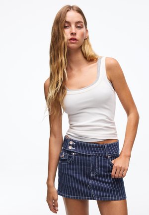 Ung kvinde med langt blondt hår iført en hvid tanktop og en blå mini-nederdel med striber, poserer mod en ensfarvet hvid baggrund.