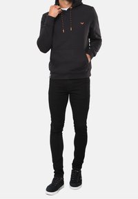 Una persona está de pie vestida con una sudadera negra con cordones naranjas, jeans skinny negros y zapatillas negras, con un fondo minimalista.
