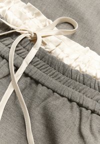 Graue Stoffshorts mit einem sanft gerafften Bund, versehen mit cremefarbenen Rüschendetails und einem cremefarbenen Kordelzug. Die Textur wirkt glatt und leicht.