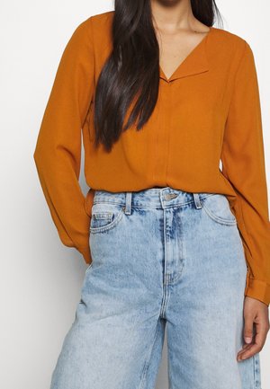 Femme portant un chemisier à manches longues orange rouille rentré dans un jean taille haute à jambes larges, délavé clair, avec poches avant et passants pour ceinture.