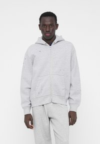 Hoodie gris zippé en mélange de coton doux. Comprend une capuche avec cordon de serrage, des poches avant et de petites éclaboussures de peinture sur le tissu.