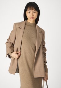 Blazer marrone con finitura testurizzata, indossato sopra un vestito a collo alto a coste dello stesso colore. Lo stile presenta ampie rever e maniche ampie.