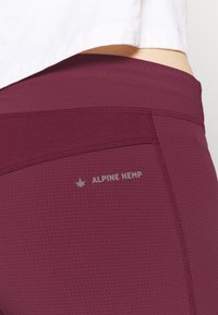Burgundy yogabyxor gjorda av en blandning av hampa, med en texturerad yta, sydda sömmar och en reflekterande "ALPINE HEMP" logotypdetalj.