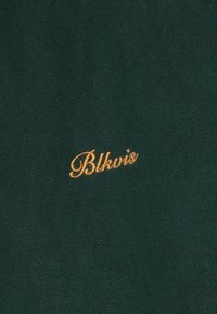 Σκούρο πράσινο ύφασμα με μικρή, καλλιγραφική πορτοκαλί κέντημα που γράφει "Blkvis" κεντραρισμένο στο ύφασμα.