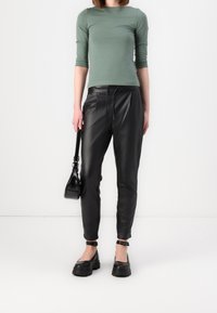 Maglia verde a maniche lunghe, pantaloni neri in pelle a vita alta con pieghe e scarpe nere con tacco spesso. Piccola borsa nera con tracolla.