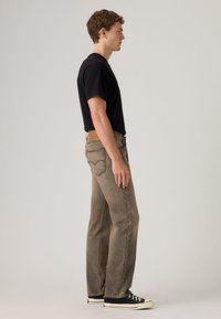 Νέος άνδρας στέκεται πλαγίως, φοράει μαύρο μπλουζάκι, ξεθωριασμένο καφέ τζιν με ίσια παντελόνια και μαύρα χαμηλά αθλητικά παπούτσια με λευκές σόλες.