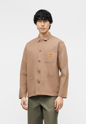 Hombre que lleva una chaqueta marrón claro con botones y tres bolsillos delanteros, y pantalones verde oliva, de pie frente a un fondo liso.