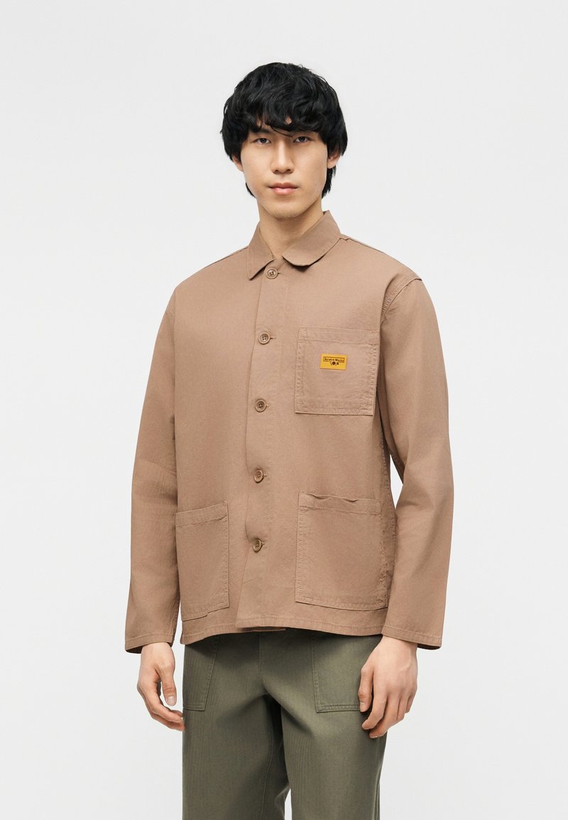 Άνδρας που φοράει ένα ανοιχτό καφέ σακάκι με κουμπιά και τσέπες μπροστά, καθώς και ελαιοπράσινο παντελόνι, στέκεται μπροστά σε απλό φόντο.