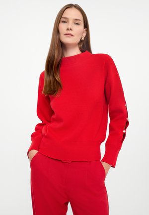 Maglione rosso lavorato a maglia con colletto alto e maniche lunghe, caratterizzato da dettagli con bottoni lungo i polsini. Texture liscia, design aderente.