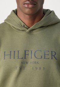 Felpa verde oliva con tasca a marsupio, caratterizzata da un grande logo blu navy che recita "HILFIGER NEW YORK EST. 1985." Tessuto in cotone morbido con dettagli di cucitura.