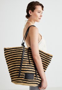 Striped woven tote bag in black and tan with leather straps and a patch label. Spacious design with a textured finish.