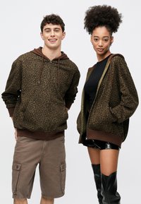 Sudaderas con capucha con cremallera de estampado de leopardo marrón y cordones, con puños y bajos acanalados. El modelo lleva pantalones cortos y otro con botas altas negras.