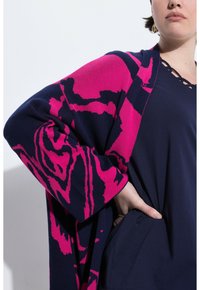 Svetr s odvážným abstraktním vzorem v růžové a námořnické modři, obohacený dlouhými rukávy a volným střihem, vyrobený z měkkého pleteného materiálu.