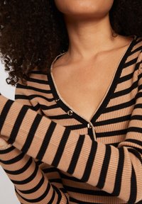 Cardigan en maille rayée beige et noir avec texture côtelée, présentant un col en V profond, une fermeture à boutons et des accents métalliques.