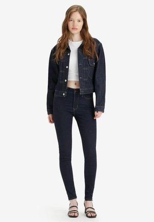 Dunkelblaue Jeansjacke mit Fronttaschen, kombiniert mit hochgeschnittenen, dunklen Skinny-Jeans und einem weißen Cropped-Top. Schwarze, geschlossene Sandalen runden den Look ab.