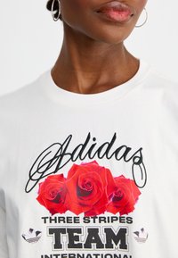 Hvid bomulds t-shirt med et blomsterdesign med røde roser, "Adidas" i kursiv og "THREE STRIPES TEAM INTERNATIONAL" i fed sort tekst.