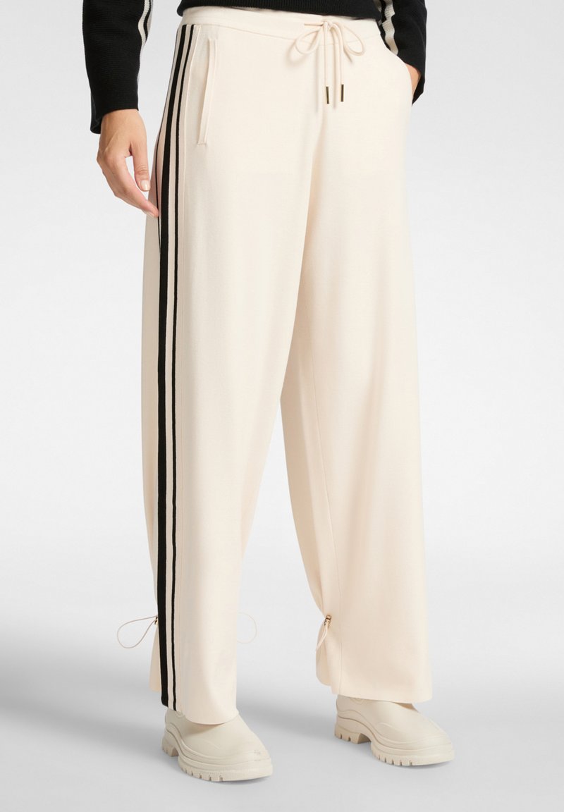 Pantalones de pierna ancha en color crema, hechos de una tela suave. Presentan franjas laterales negras, una cinturilla con cordón y cremalleras en los tobillos.