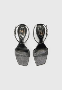 Μαύρα γκλιτερ πέδιλα με λουράκι στο πίσω μέρος, τετράγωνη μύτη και σχεδιασμό με σταυρωτά λουράκια. Διαθέτουν μαύρα εσωτερικά πέλματα με μεταλλικό λογότυπο.