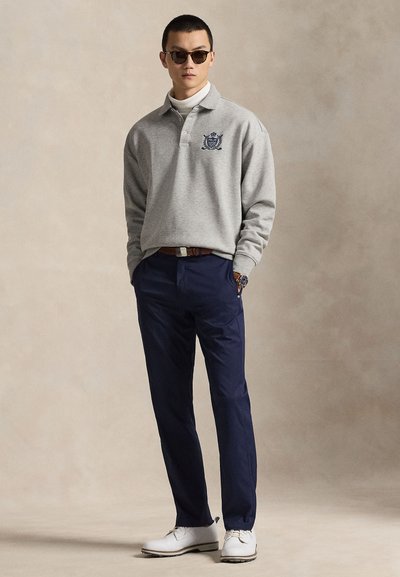 Γκρι μακρυμάνικη πόλο μπλούζα με κεντημένο οικόσημο, συνδυασμένη με ναυτικό μπλε παντελόνι. Αξεσουάρ περιλαμβάνουν σκούρα γυαλιά ηλίου και λευκά παπούτσια.