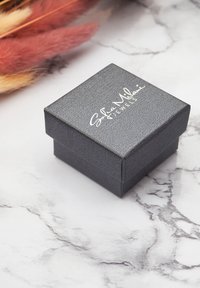 Svart texturerad smyckeslåda i fyrkantig form, med silverfärgad märkning "Sofia Milani Jewels" på locket, placerad på en marmor yta.