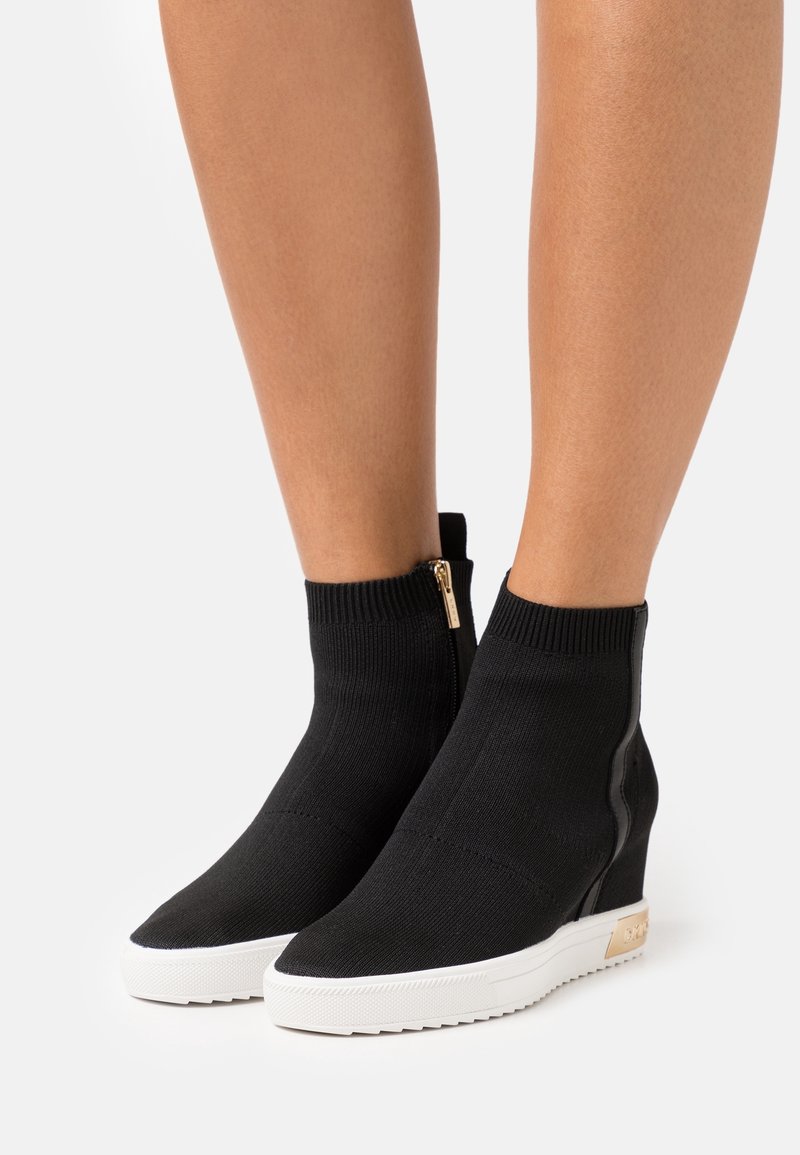 Bottines en maille noire avec semelles plateforme blanches, fermeture éclair dorée sur le côté et talons compensés, portées sur des jambes nues.
