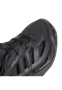 Primo piano di una scarpa da ginnastica nera che mostra pannelli laterali in mesh testurizzato, sovrapposizioni sintetiche modellate e lacci neri su sfondo bianco.