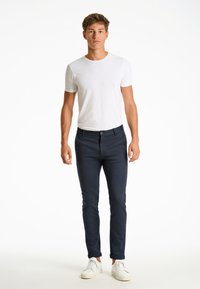 Navyblaue Slim-Fit-Hose mit Stoffstruktur, ausgestattet mit einem vorderen Knopfverschluss, kombiniert mit einem schlichten weißen T-Shirt mit kurzen Ärmeln und weißen Sneakers.