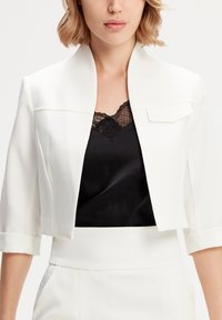 Veste blanche cropped avec un col montant et des manches courtes, présentant une texture lisse et une poche avant unique. Débardeur en dentelle noire en dessous.