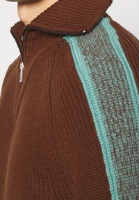 Närbild av en brun stickad tröja med dragkedja i kragen och en vertikal turkos stickad rand längs axeln.