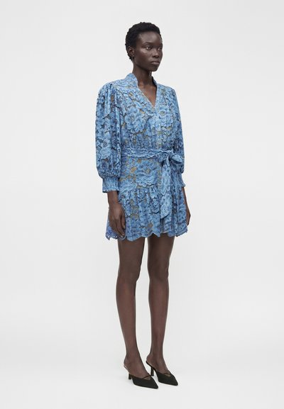 Femme debout portant une robe en dentelle bleue à motifs floraux, avec des manches longues bouffantes, une ceinture à la taille et des talons noirs pointus sur un fond clair uni.