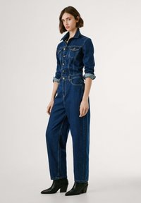 Jeans jumpsuit med lange ærmer, knappestykke foran og rullede mansjetter. Mørk blå farge, smale ben og en figurnær midje. Svart hælstøvler.