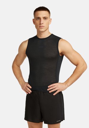 Atletyczny mężczyzna stojący z rękami na biodrach, ubrany w czarny bezrękawnik z siateczki i czarne sportowe spodenki na jednolitym tle.