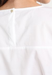 Blusa bianca vista dal retro con scollatura tonda, caratterizzata da un piccolo bottone e dettagli di cucitura, realizzata in tessuto leggero.