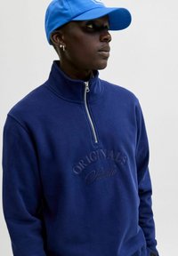 Felpa zip con colletto alto di colore blu navy, texture morbida e testo ricamato "ORIGINALS". Indossata con un cappellino blu.