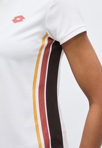 Λευκό πουκάμισο με κοντά μανίκια, με κατακόρυφες ρίγες σε μούστο, σκουροκόκκινο και μαύρο κατά μήκος της πλευράς και ένα μικρό κόκκινο λογότυπο κοντά στον ώμο.