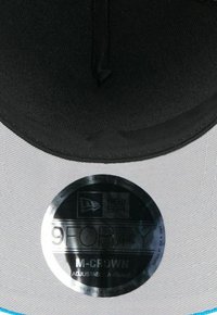 Schwarz-graue Mütze mit flachem Schild, ausgestattet mit einem glänzenden runden Etikett, das "9FORTY M-CROWN Adjustable A-Frame" anzeigt, und strukturiertem Stoff.