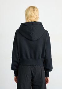 Haut à capuche noir court avec une capuche spacieuse, des poignets côtelés et une texture douce. Porté avec un pantalon noir ample, affichant un design minimaliste.