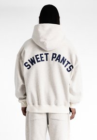 Haut à capuche gris clair avec un grand texte navy "SWEET PANTS" au dos, doté d'une capuche avec cordon de serrage et de poignets et ourlet côtelés. Texture douce.