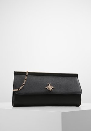 Musta suorakaiteen muotoinen clutch-laukku, jossa on teksturoitu läppä, kultainen mehiläiskoriste ja kultainen ketjuhihna, lepää valkoisella pinnalla.