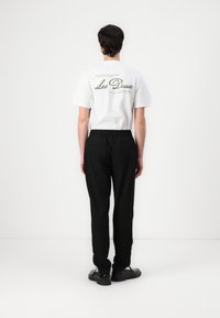 White cotton t-shirt with green text on back, paired with black trousers, worn by a person facing away, with black footwear and elastic waistband.
