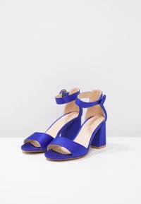 Pár královsky modrých saténových sandálů na blokovém podpatku s pásky kolem kotníků a otevřenou špičkou, vystavených na bílé ploše a světle šedém pozadí.