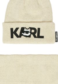 Gorro de punto beige con puño doblado, que presenta letras "KARL" en negro y un gráfico de un gato blanco con gafas de sol. Textura suave y diseño minimalista.