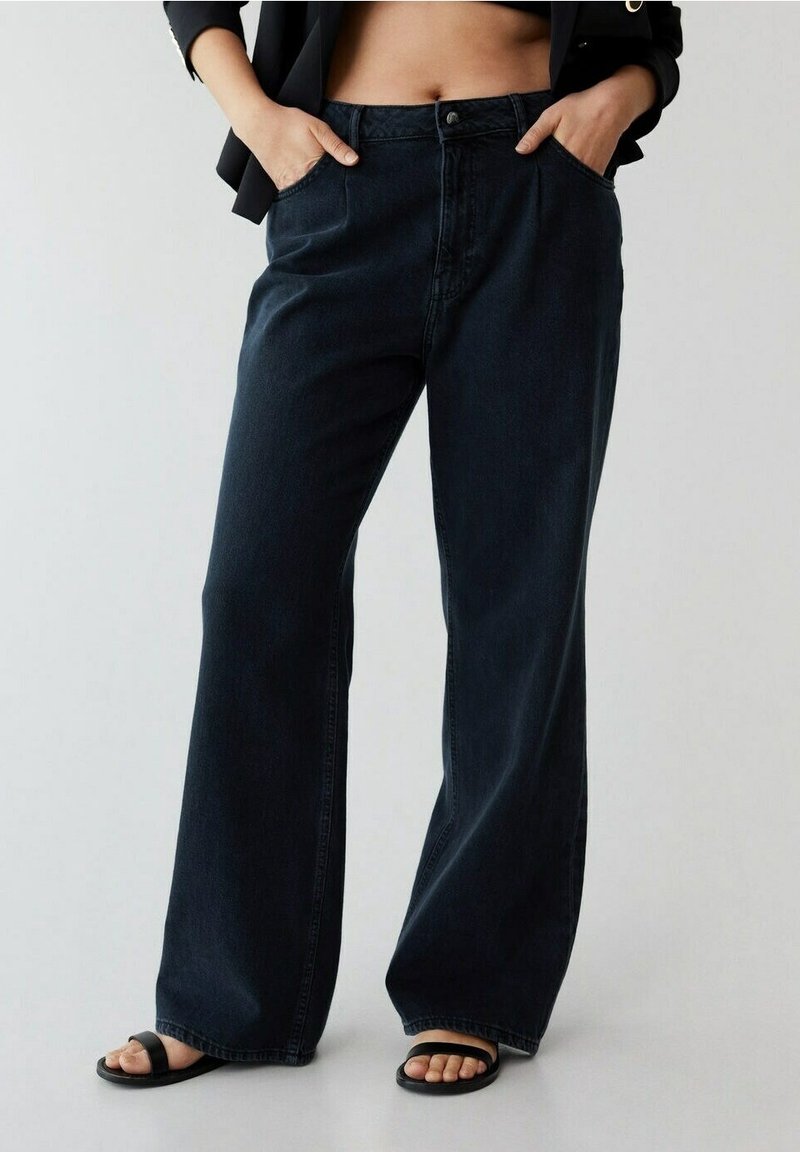 Personne portant un jean noir taille haute à jambe large, les mains dans les poches et des sandales noires à bout ouvert, debout devant un fond uni.