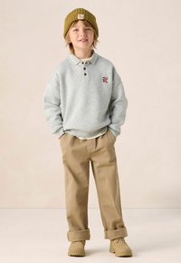 Grauer Sweatshirt mit Kragen und Knöpfen, beige Hose, die an den Bündchen aufgerollt ist, und einer gerippten olivgrünen Mütze. Mit einem kleinen farbenfrohen Aufnäher.