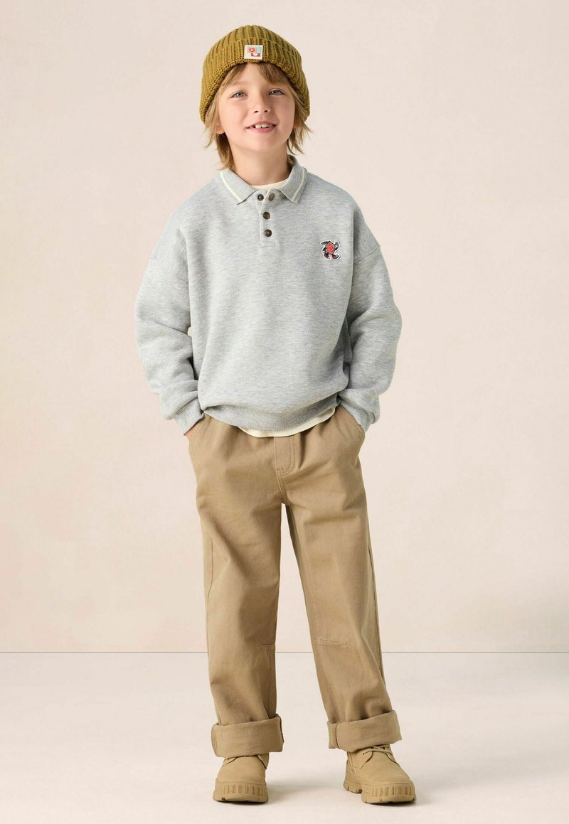 Grauer Sweatshirt mit Kragen und Knöpfen, beige Hose, die an den Bündchen aufgerollt ist, und einer gerippten olivgrünen Mütze. Mit einem kleinen farbenfrohen Aufnäher.