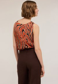 Top senza maniche colorato con un motivo astratto arancione e marrone; abbinato a pantaloni marroni a vita alta, che mettono in mostra una texture liscia.
